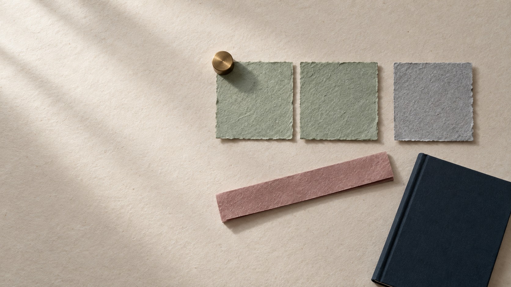 Editorial still life of three squares of handmade paper in a row, two celadon green and a third grey one set slightly apart, on warm textured paper