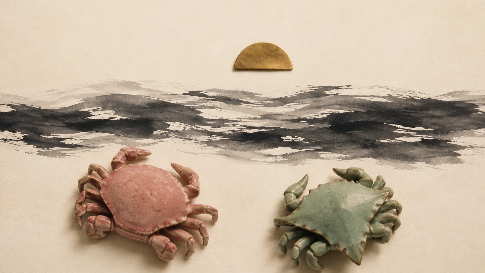 Editorial still life of two ceramic crab figures, one rose with a rounded shell and one celadon with a pointed shell, separated by a band of dark ink brushwork