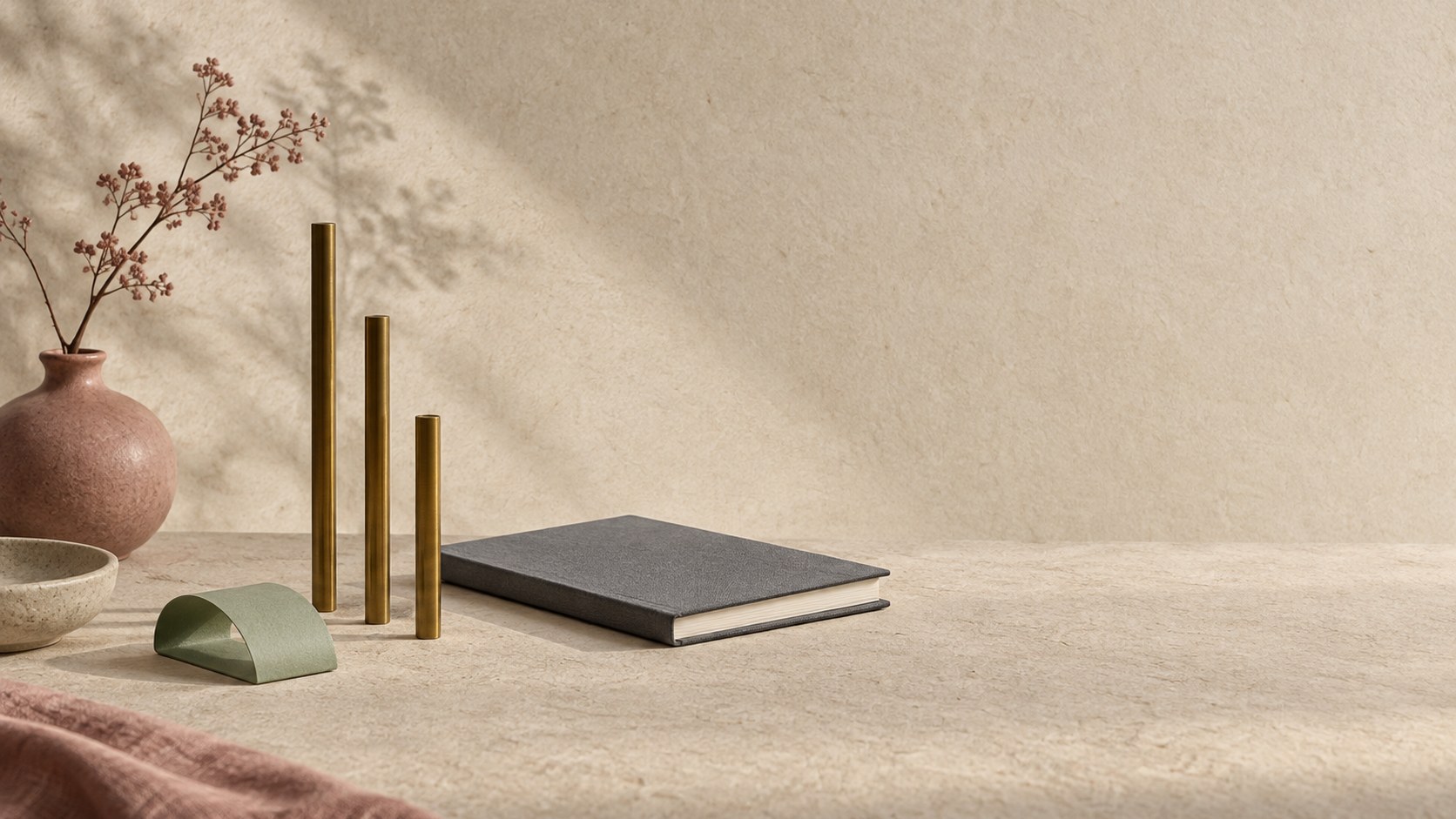 Editorial still life of three brass rods of descending height standing beside a folded celadon paper band and a closed grey notebook on warm textured paper