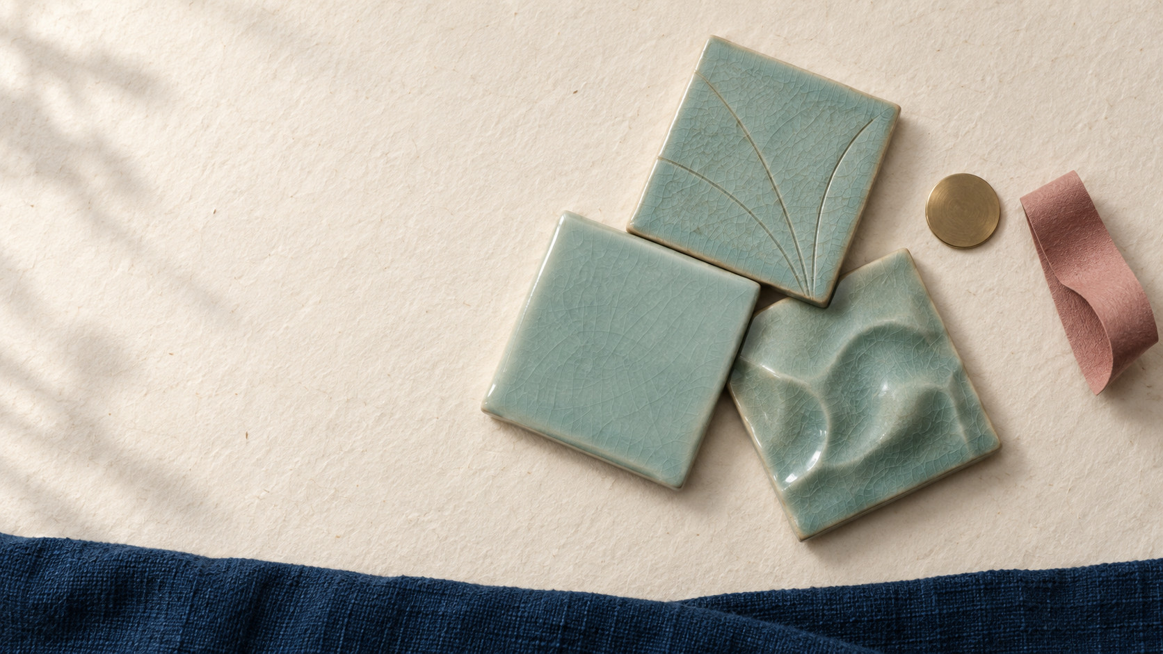 AI-generated editorial still life of three celadon-glazed tiles, one carved, one smooth and one raised, touching at their corners beside a brass disc