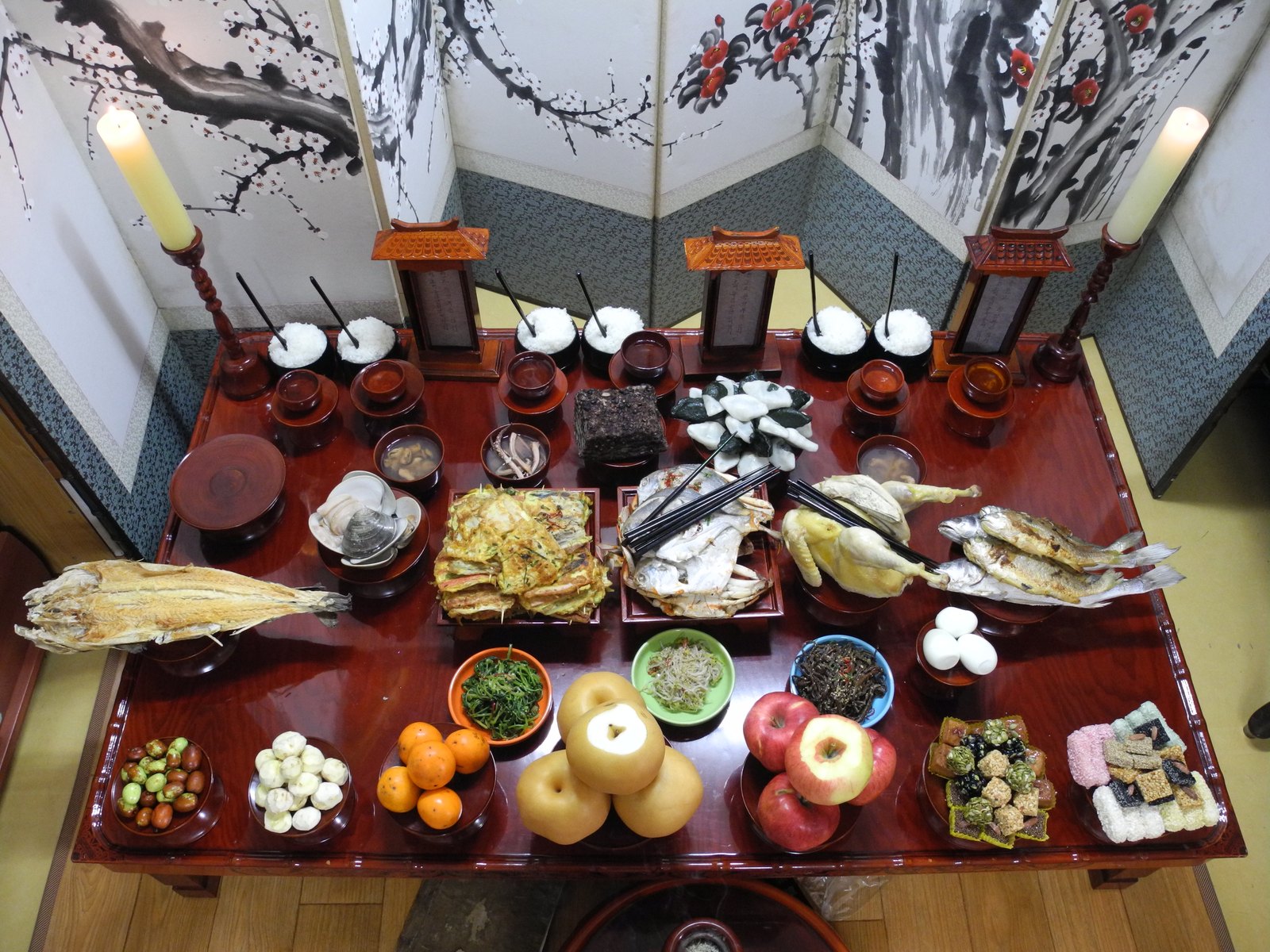 A formally set Chuseok charye table seen from above, with rice bowls, fish, fruit and rice cakes laid out in rows in front of a folding screen