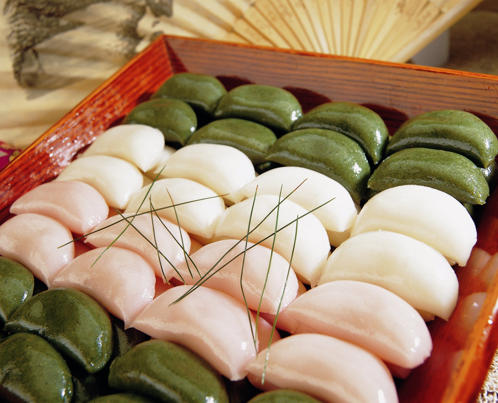 Rows of pink, white and green songpyeon rice cakes arranged in a red lacquered tray with pine needles laid across them