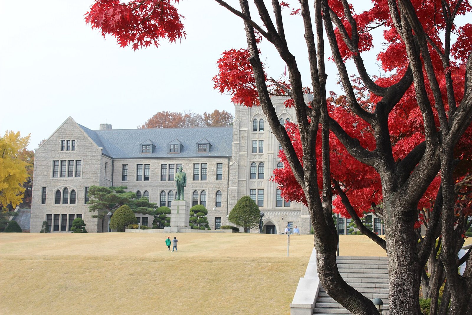 Korea University's granite main building and statue across an autumn lawn on the Anam campus, framed by a red maple, two tiny distant figures on the grass
