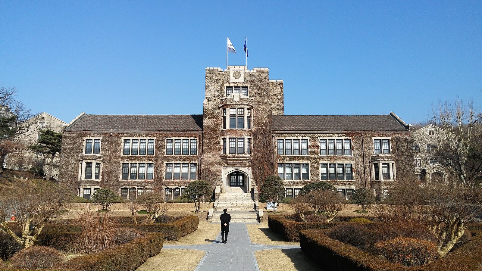 A stone Gothic-style hall with a central tower and flags on Yonsei University's Sinchon campus, a single distant figure walking away along a garden path