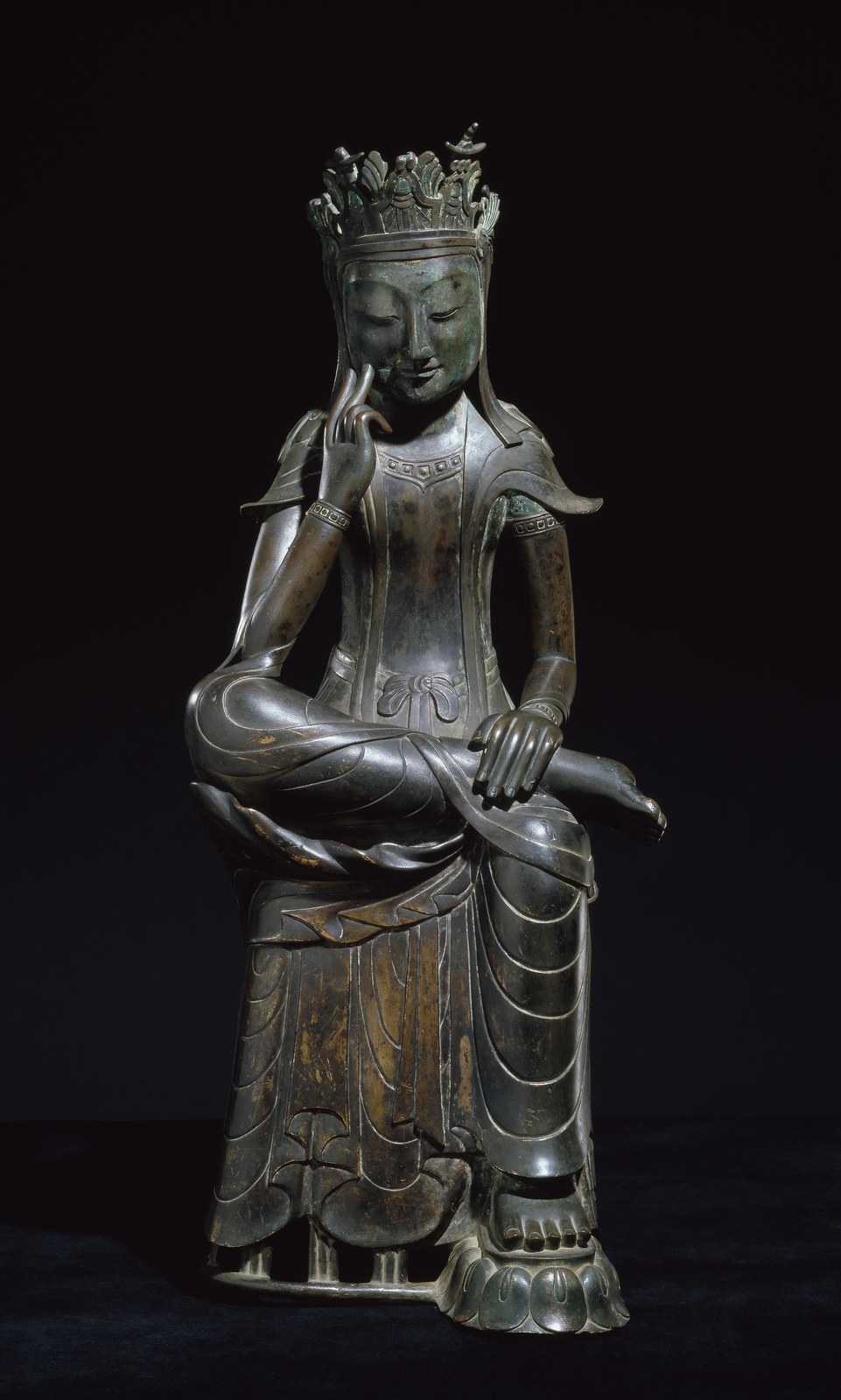 Gilt-bronze pensive bodhisattva statue seated with one ankle over the knee and fingers touching its cheek, photographed against a black background