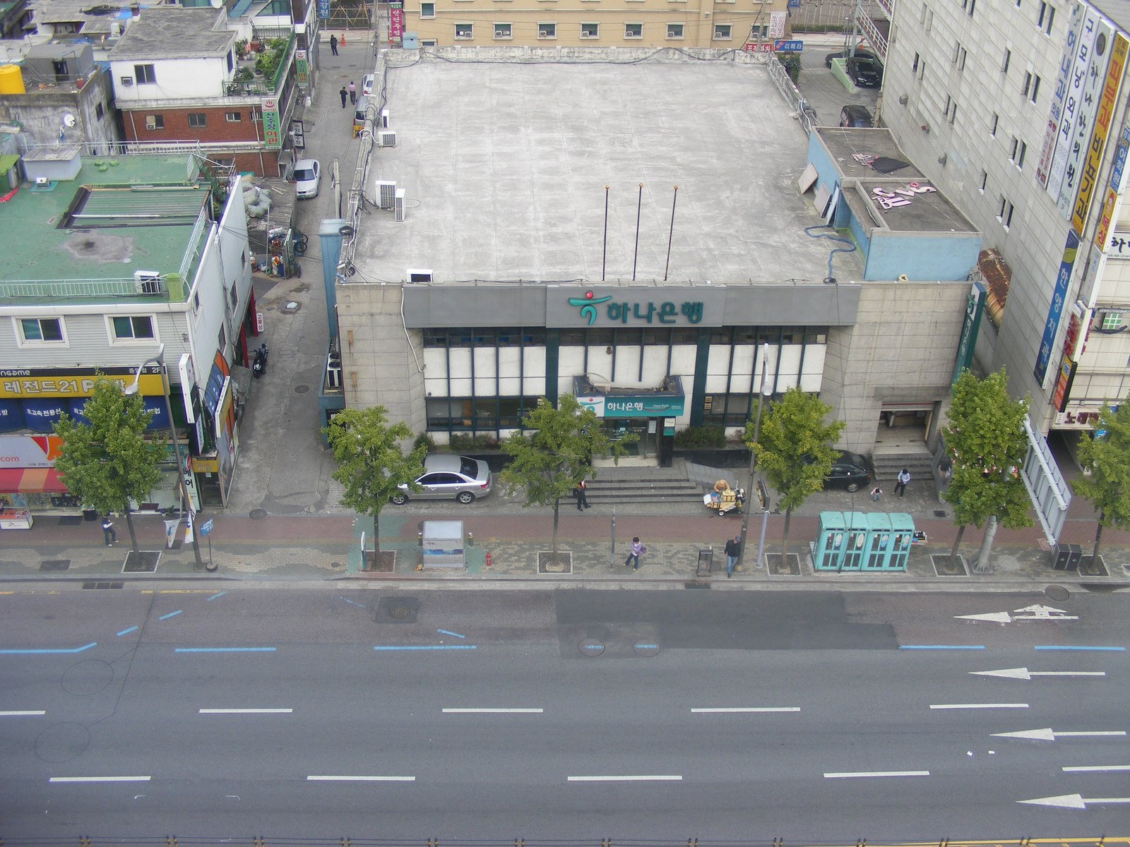 Aerial view of a stand-alone two-storey Hana Bank branch building on a wide multi-lane city street, photographed from a tall building across the road