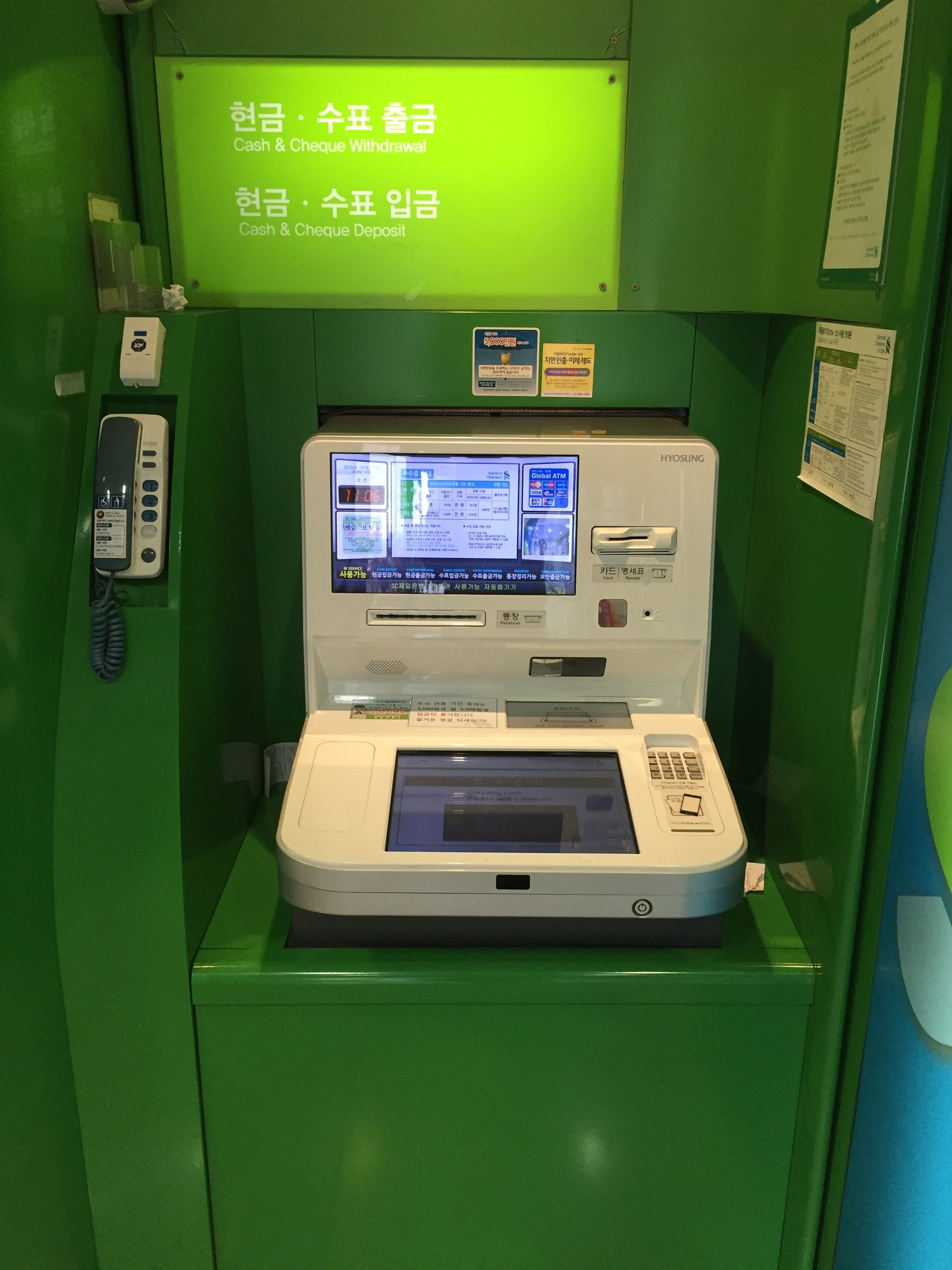 Green self-service ATM booth in Seoul with a Hyosung machine, bilingual cash and cheque withdrawal signs and a courtesy phone on the wall