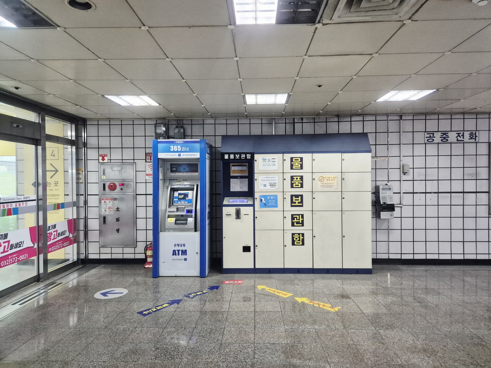A blue shared-network ATM kiosk beside a wall of parcel lockers and a payphone in a tiled Incheon subway station corridor