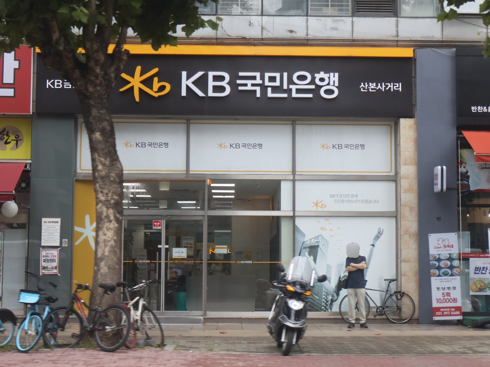 Street-level KB Kookmin Bank branch with a dark signboard and yellow trim at a city intersection, with bicycles and a scooter parked outside