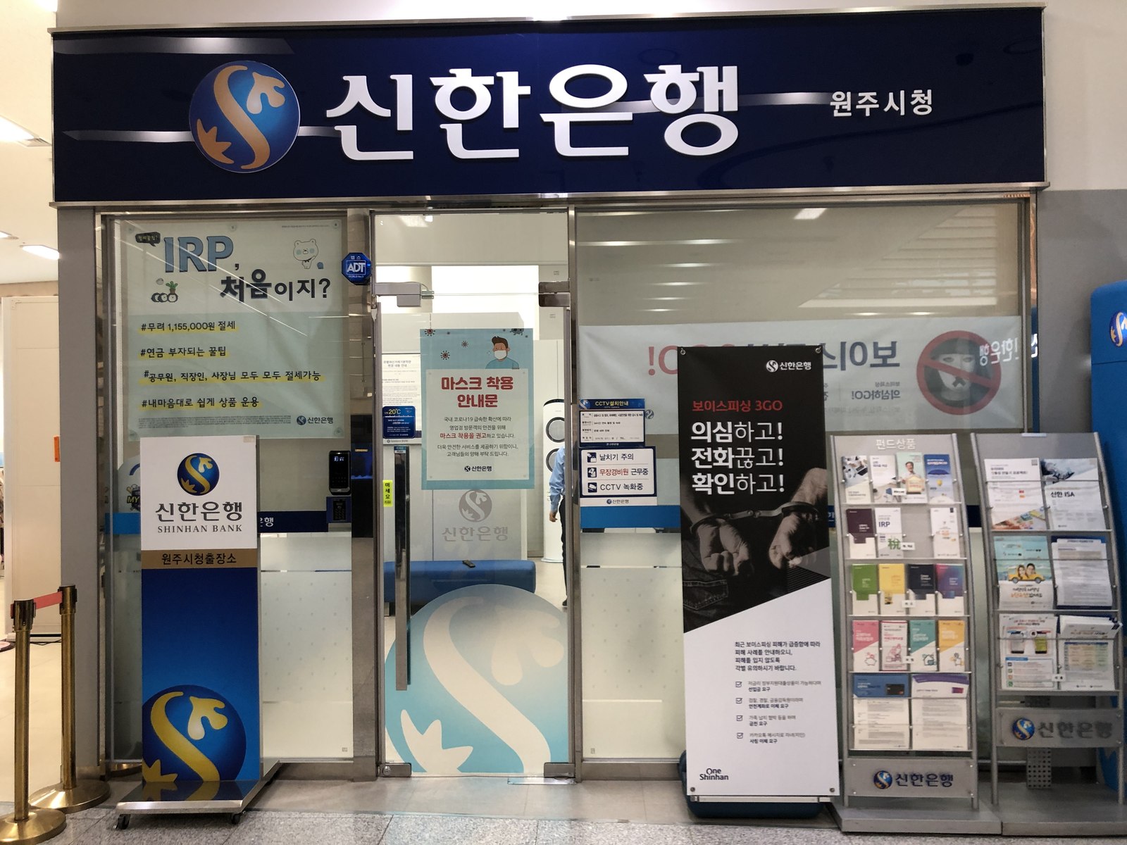 Entrance of a small Shinhan Bank service branch inside Wonju City Hall, with a tall voice-phishing warning banner standing beside the glass doors