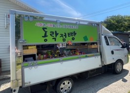 A small white mobile-market truck with a green open sales flap and shelves of goods parked beside a rural Korean road