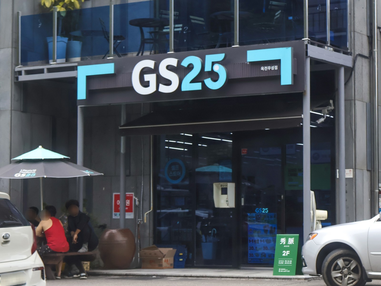 Street-level GS25 convenience store beneath a dark modern building in Okcheon town centre, with a parasol and parked cars outside