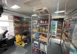 Inside a mobile market truck: metal shelving stocked with drinks, sauces and packaged foods beside glass-door fridges