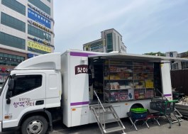 A larger mobile market truck with an open side wall and steps, stocked with packaged foods, parked on a town street