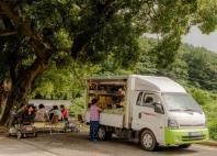 A mobile market truck with its side open, parked under a large tree while villagers browse goods in a rural Korean village
