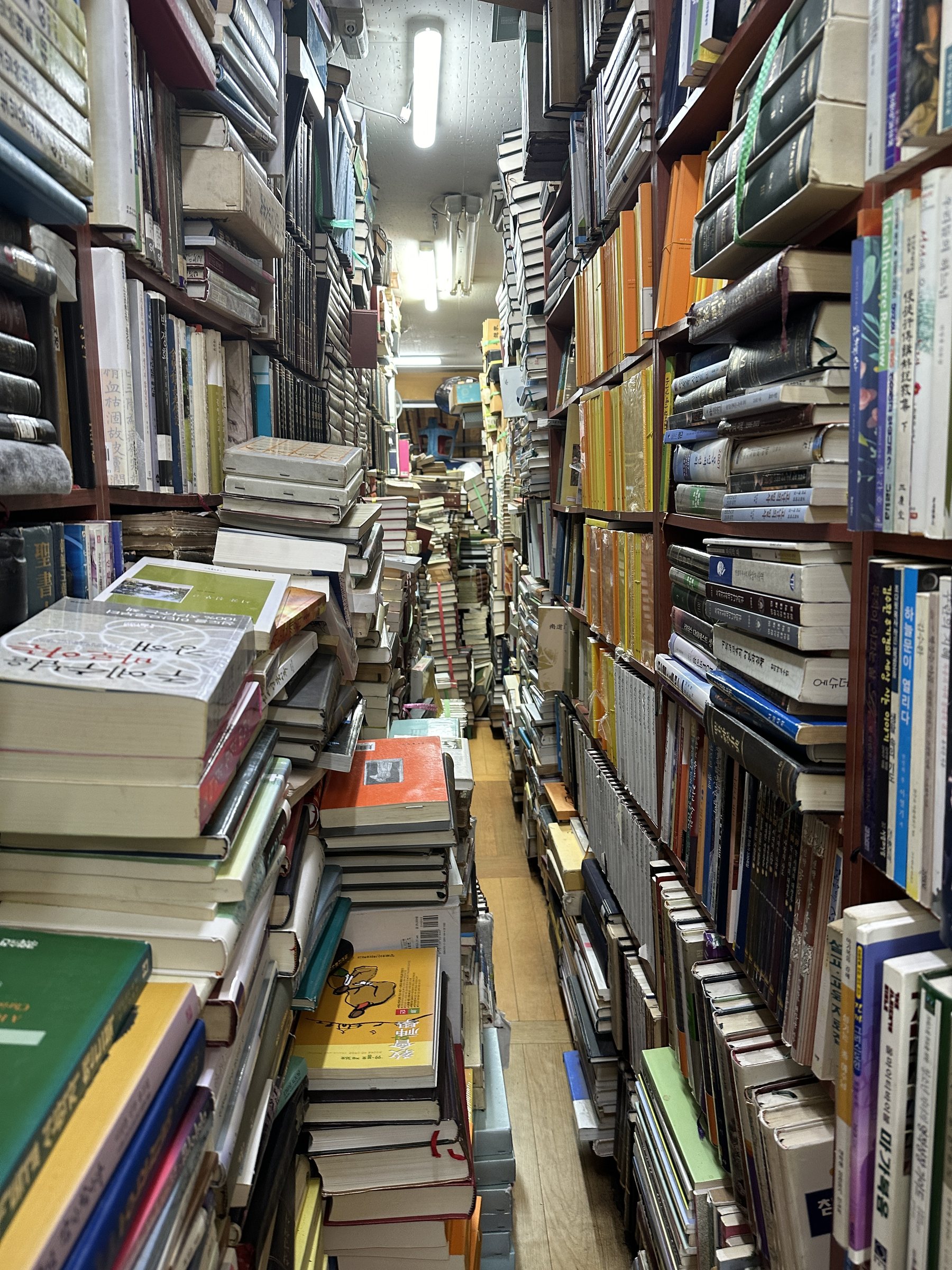 Floor-to-ceiling stacks of secondhand books rising in a narrow Seoul bookstore aisle