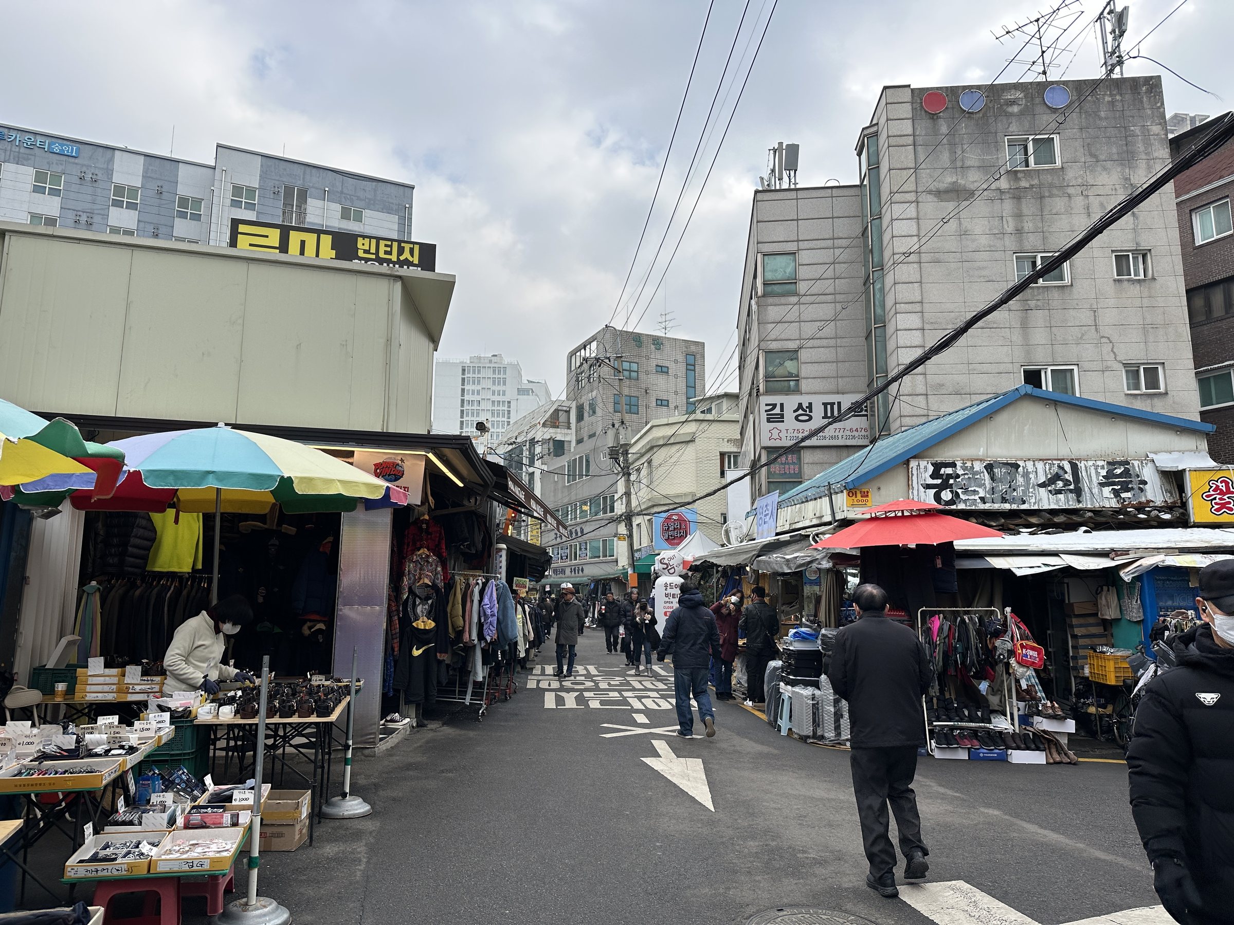 Winter street view of the Dongmyo flea market alley in Seoul, with vendors' umbrellas, clothing stalls and shoppers in coats