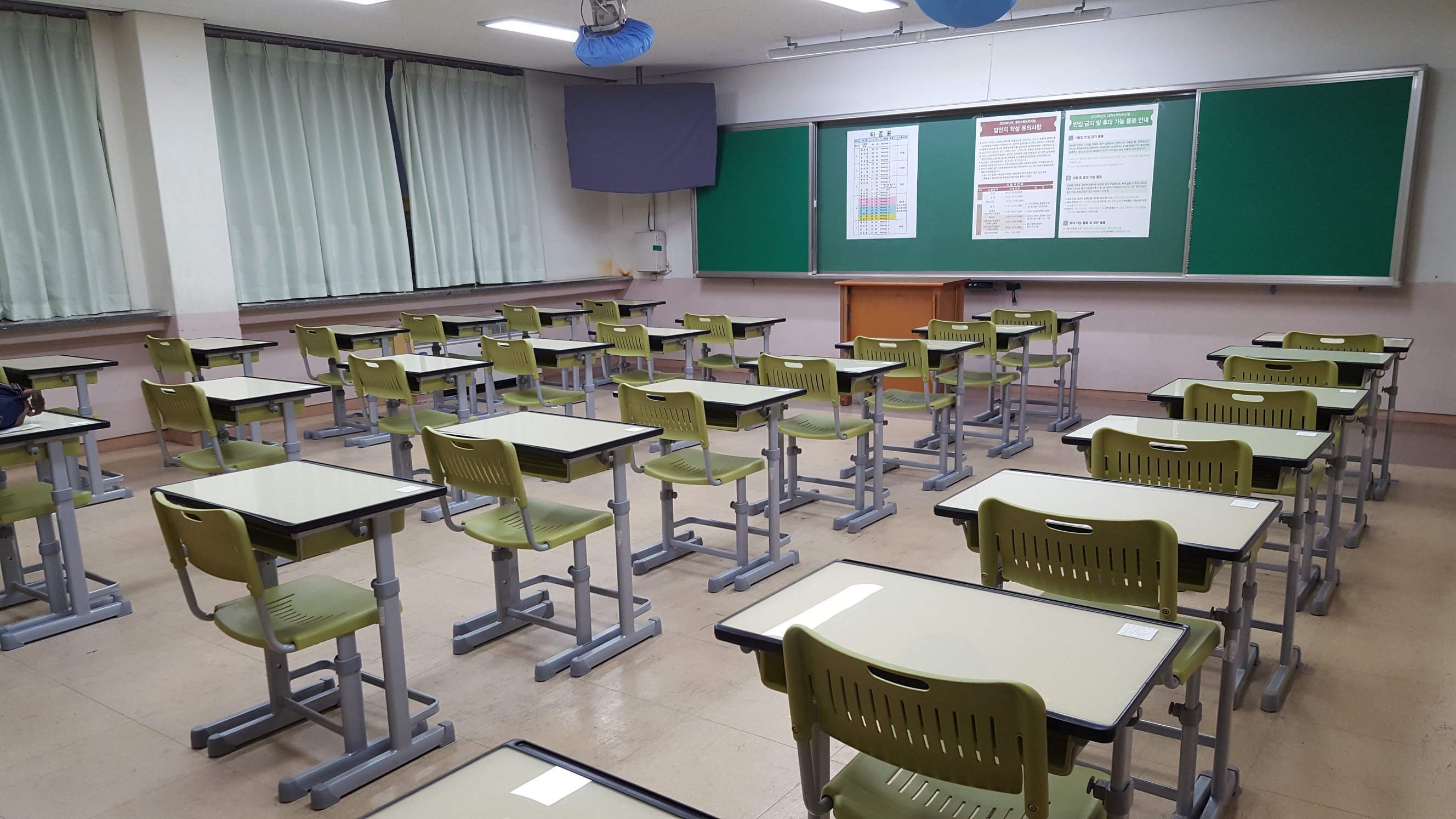 An empty Korean high-school classroom arranged as an exam room, single desks in neat rows with seat-number slips and schedule charts posted on the green chalkboard