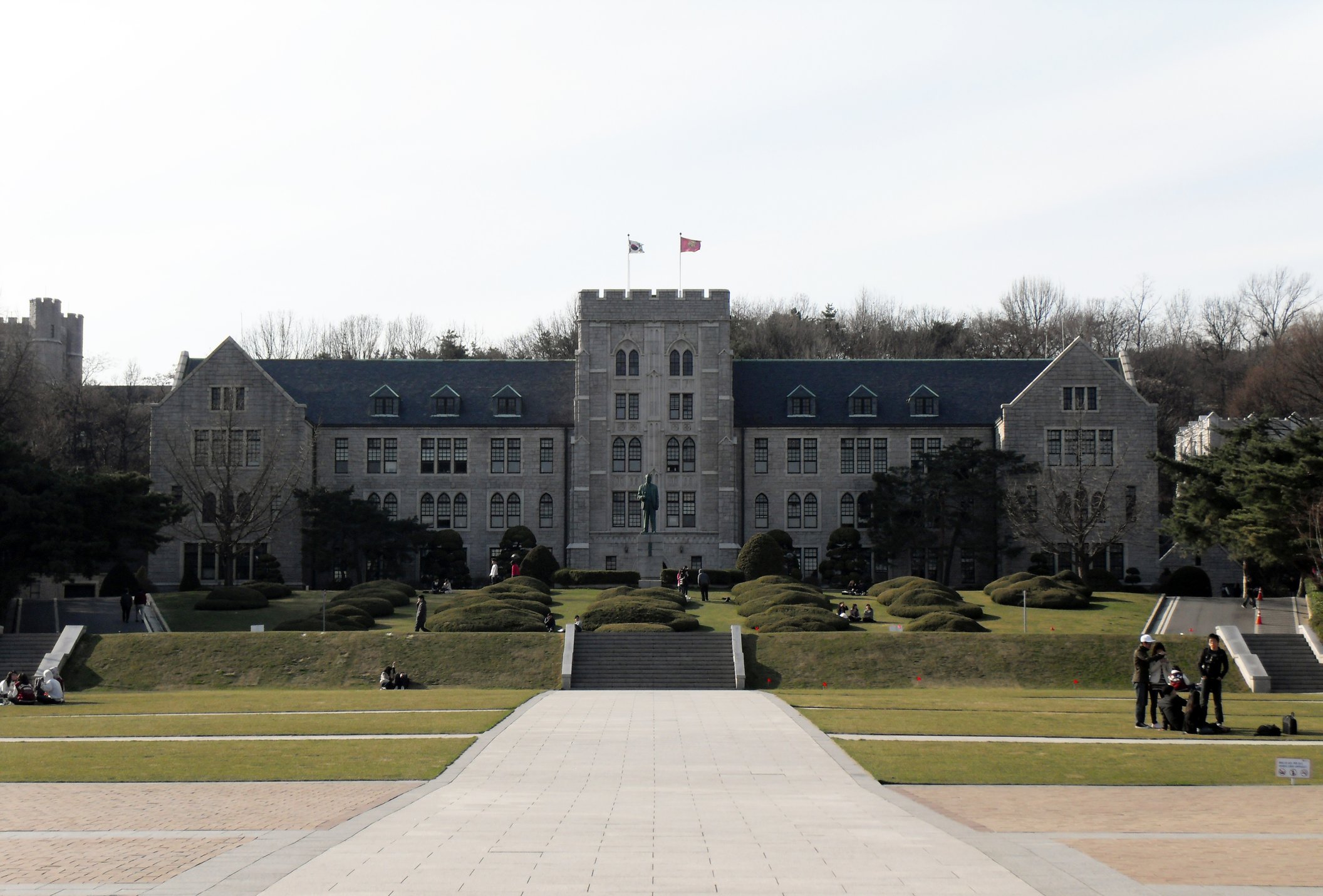 The granite Gothic-revival main hall of Korea University with its crenellated central tower, seen across a lawn plaza with a few small distant figures
