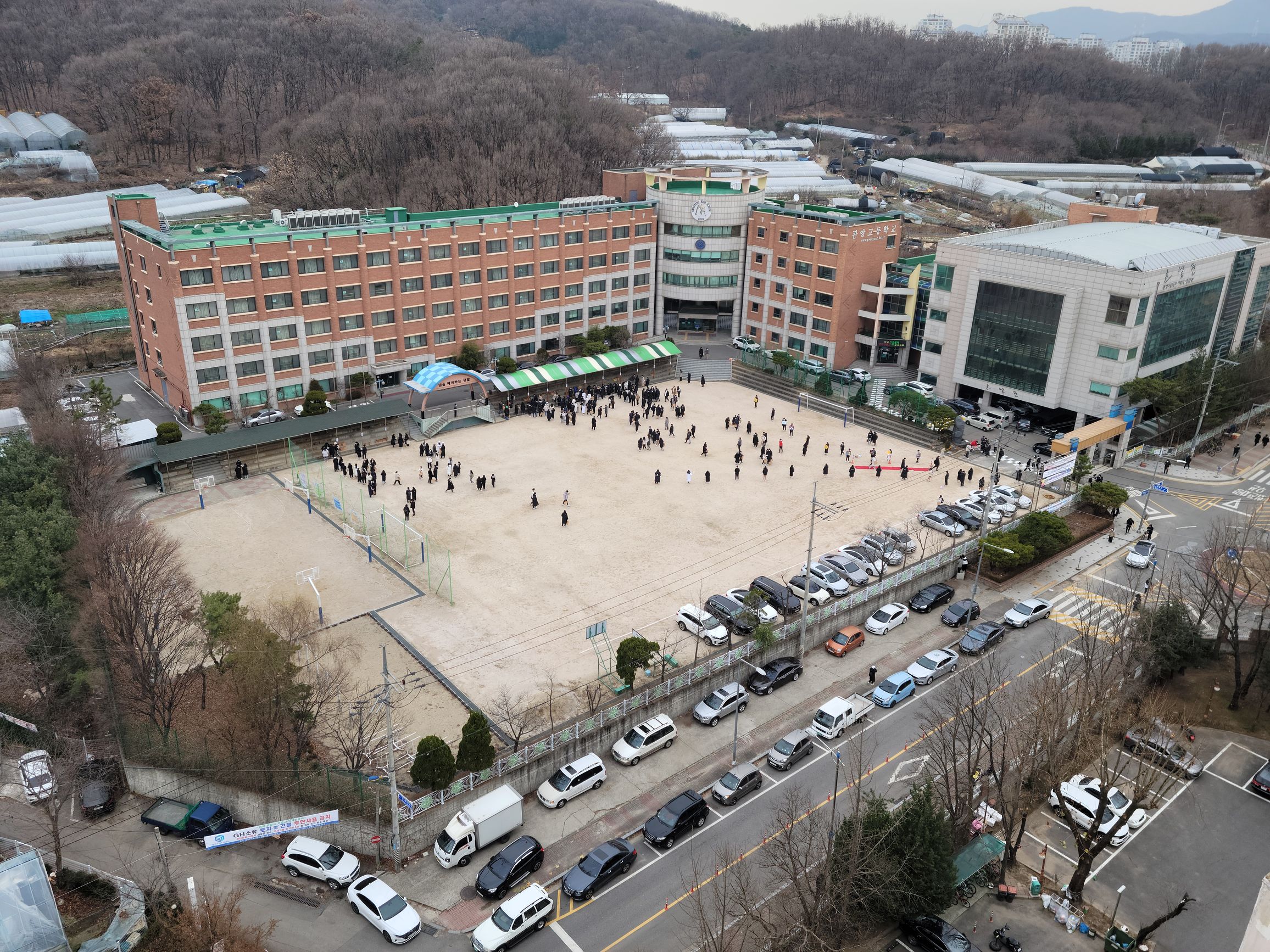 High-angle view of a school field crowded with hundreds of small distant figures gathering for the exam preliminary call-up, cars lining the street outside