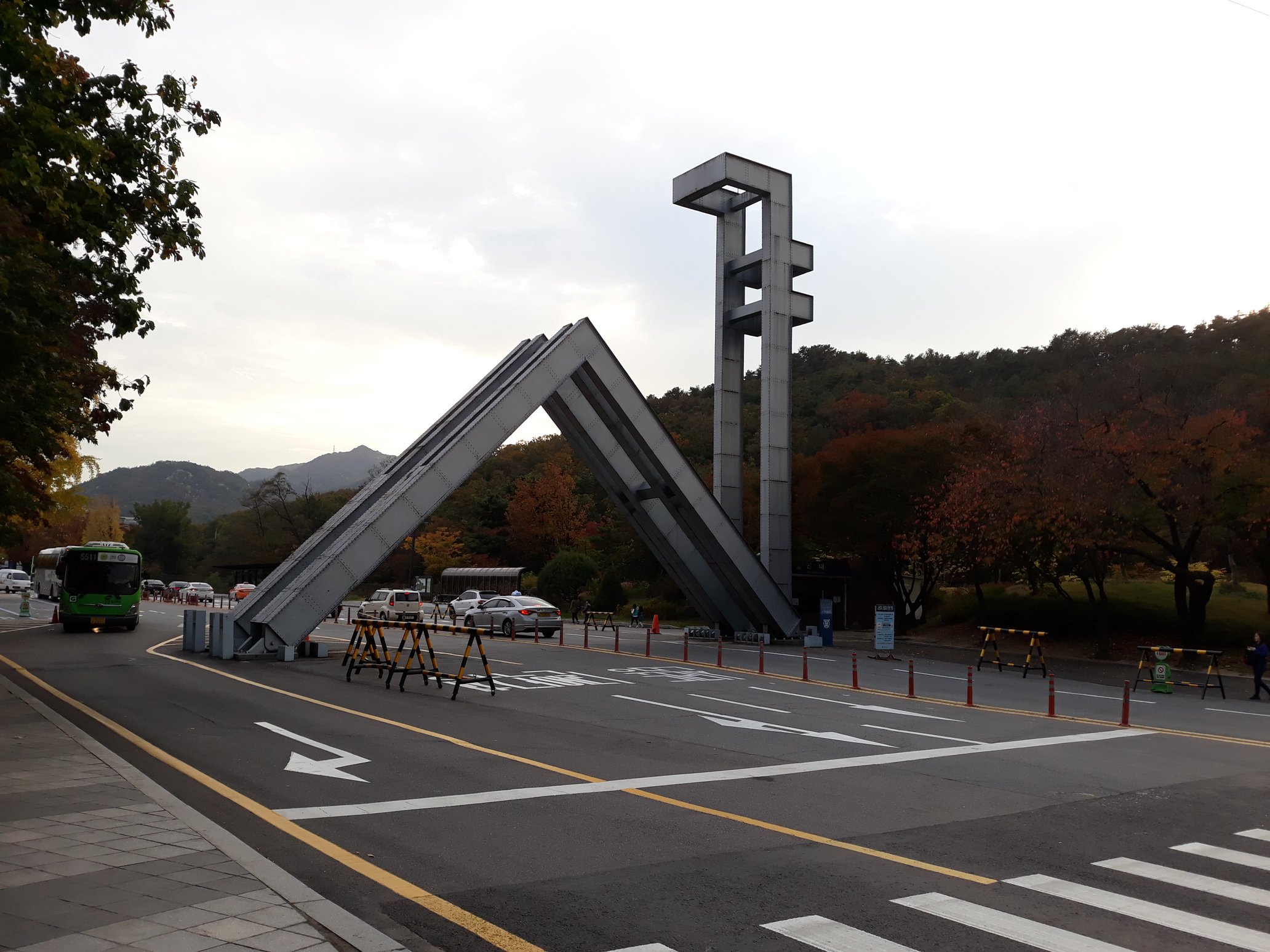 The angular concrete main gate sculpture of Seoul National University over the campus entrance road, with an autumn hillside and a green shuttle bus behind