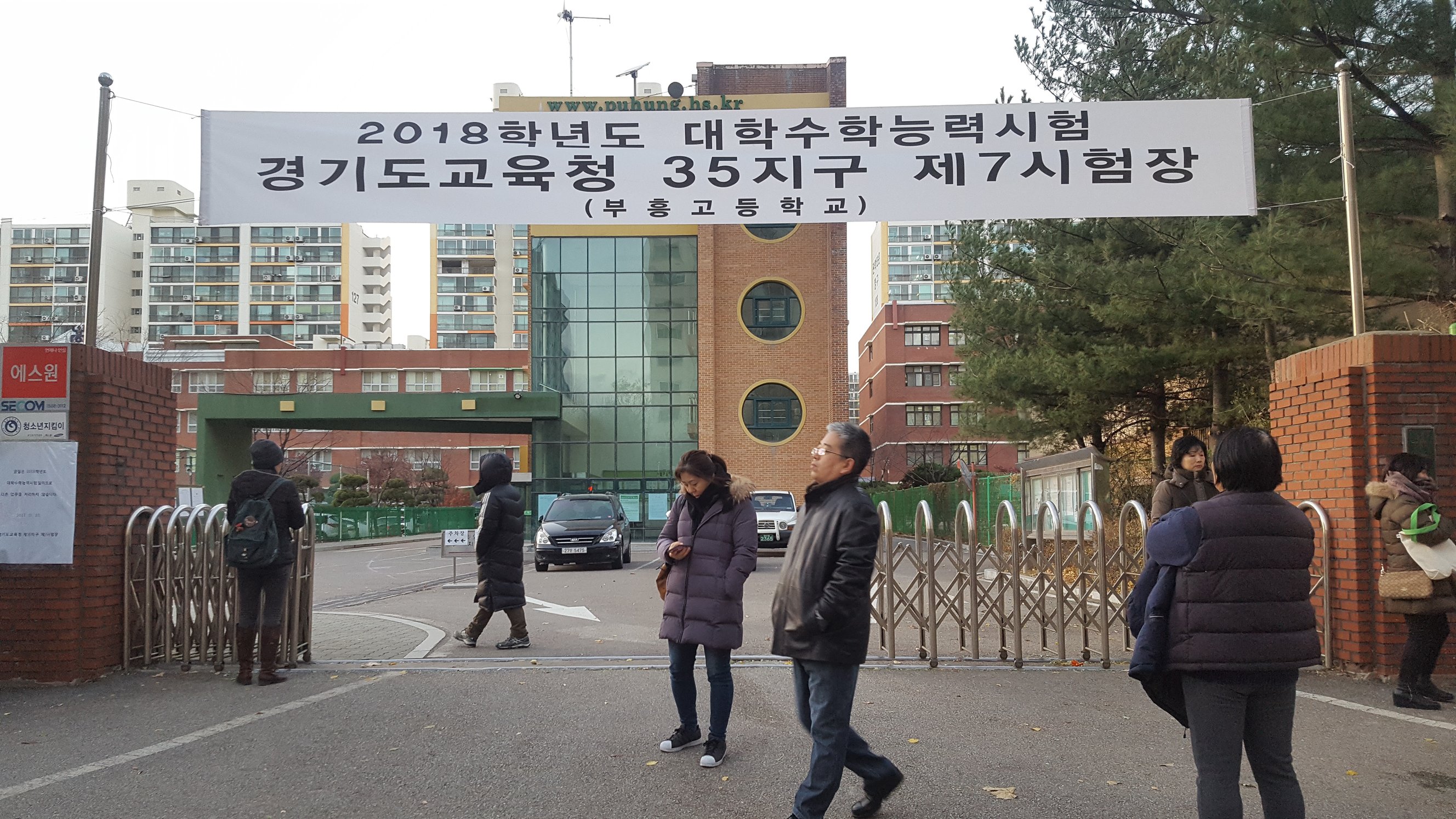 A school main gate with a large white banner marking the building as a College Scholastic Ability Test site, a few adults walking out on exam day