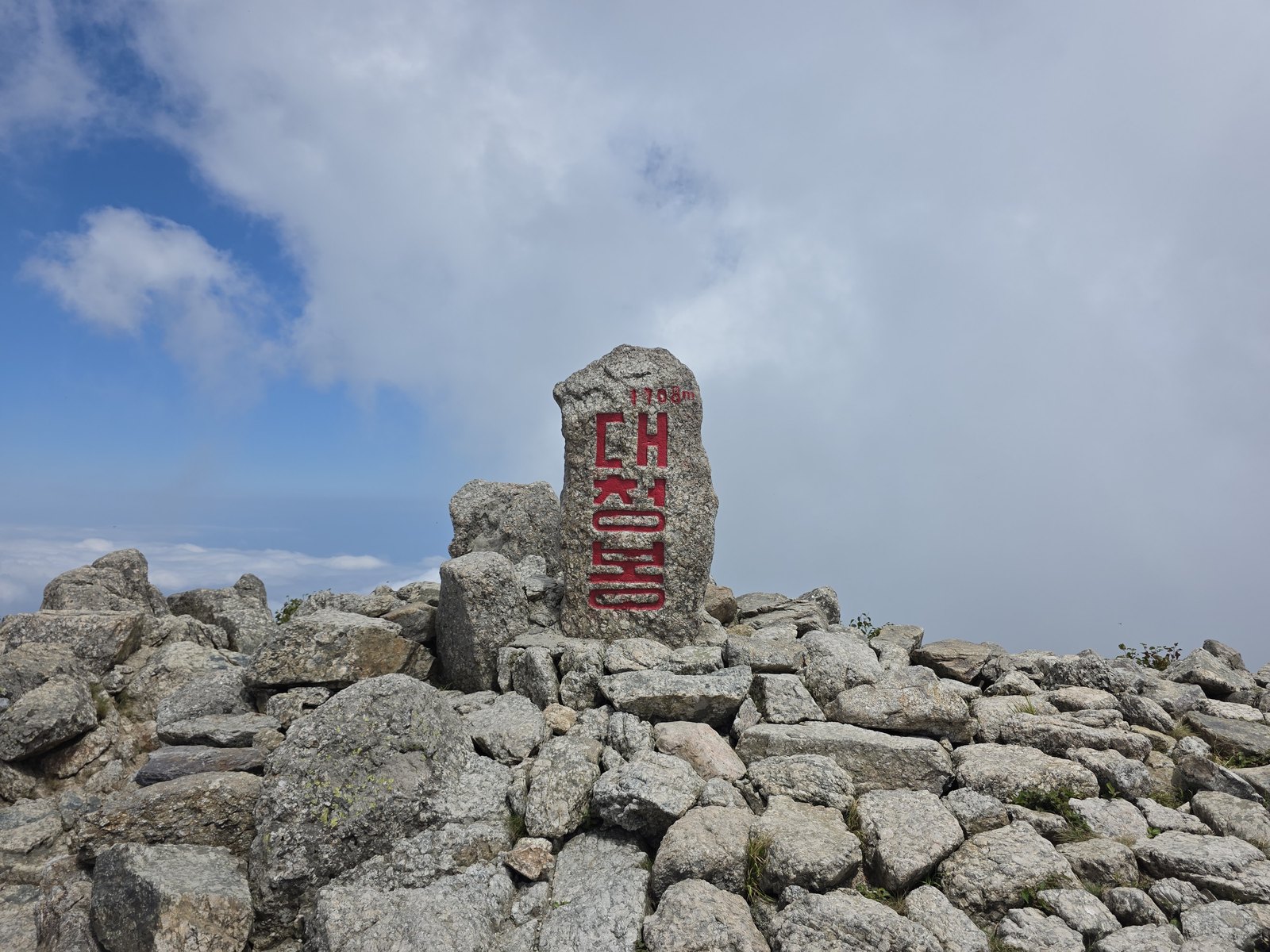 Stone summit marker on a bare rocky ridge under a pale blue sky with cloud below, standing among loose granite blocks