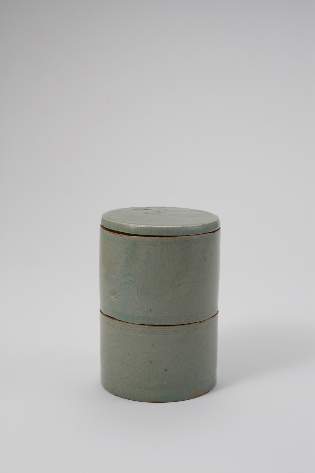 Goryeo celadon lidded box, a tall two-tier grey-green cylinder with a flat lid, faint fine lines between narrow patterned bands on its side and a crackled glaze, on a plain grey background