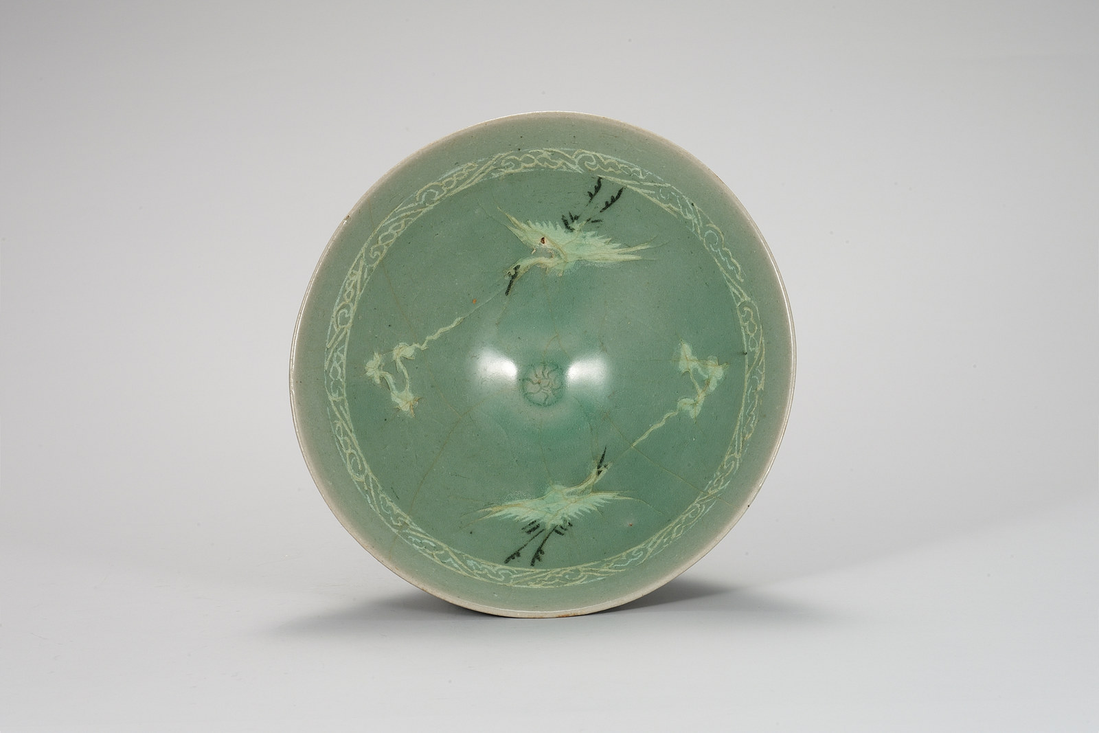 View into a Goryeo celadon bowl from above showing two pale cranes with dark details and two pale cloud motifs on green glaze, ringed by a pale scroll band near the rim